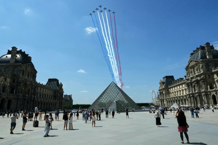 Des passants regardent la Patrouille de France (PAF) survoler la pyramide du Louvre conçue par l'architecte sino-américain Ieoh Ming Pei, le 9 juillet 2025 à Paris, lors d'une répétition du défilé annuel du 14 juillet