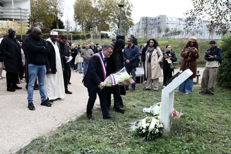 Le frère de Bouna, Siyakha (g), et son père Seydou (2e g) debout près du maire de Clichy-sous-Bois, Olivier Klein (c), qui dépose des fleurs devant une plaque à l'extérieur du lycée Robert Doisneau lors d'une cérémonie  marquant le 20e anniversaire de la mort des adolescents Zyed Benna et Bouna Traoré, le 27 octobre 2025 en Seine-Saint-Denis