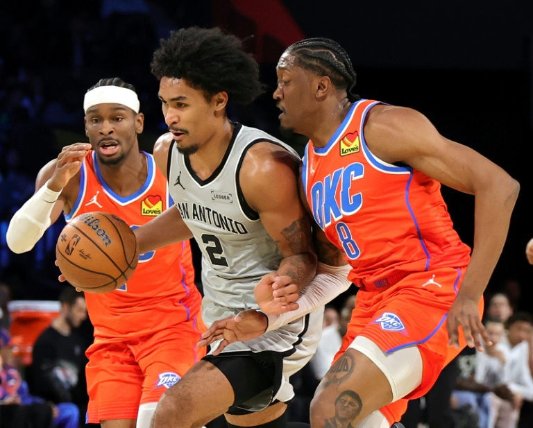 Dylan Harper (c) s'inflitre entre Shai Gilgeous-Alexander (g) et Jalen Williams lors du match de NBA entre les San Antonio Spurs et l'Oklahoma City Thunder, le 13 décembre 2025 à Las Vegas