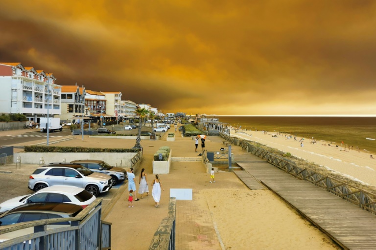 Des vacanciers marchent le long de la plage de Lacanau-Océan, dans le sud-ouest de la France, le 24 juillet 2026, tandis que d'épais nuages de fumée s'élèvent dans le ciel au-dessus d'un incendie de forêt